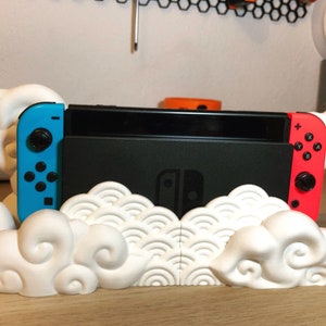 Puede incluir: Un soporte blanco impreso en 3D para una consola Nintendo Switch, con un diseño de nube. El soporte sostiene la consola negra con los mandos Joy-Con azules y rojos. El soporte está sobre una superficie de madera.
