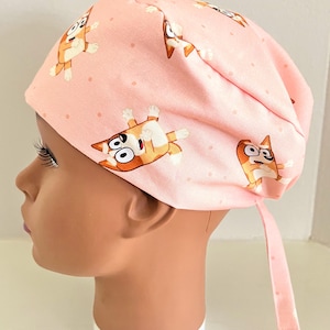May include: Pink surgical scrub hat with a cartoon dog print. The hat has a tie at the back.