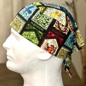 May include: A colorful, patterned head covering featuring a stained-glass design with the words "Slytherin" and "Gryffindor". The head covering is made of fabric and is tied at the back. The design includes various house crests and symbols.