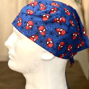 May include: A blue surgical scrub cap with a pattern of red and white Spider-Man mask designs and spiderwebs. The cap is tied at the back and is displayed on a white mannequin head.