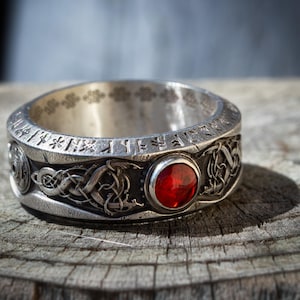 Puede incluir: Un anillo de plata con una piedra preciosa roja y grabados intrincados. El anillo presenta un diseño de nudo celta y runas alrededor de la banda.