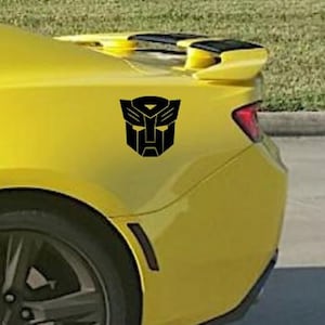 May include: A yellow car with a black Transformers logo decal on the rear.