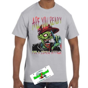 Puede incluir: Camiseta gris claro con un gráfico de zombi y el texto "ARE YOU READY FOR A ZOMBIE ATTACK". El zombi tiene piel verde y un sombrero rojo. Una placa de matrícula de Florida está en la esquina inferior izquierda.