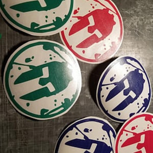 May include: A collection of circular stickers in various colors, including green, red, and blue. Each sticker features a white graphic design within a white border and a splatter effect. The stickers are arranged on a dark, textured surface.