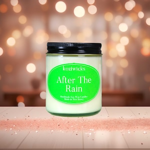 Pode incluir: Uma vela de cera de soja branca em um frasco de vidro com tampa preta. O rótulo é verde e diz "koziwicks After The Rain Handmade Soy Wax Candles Made in New Jersey".