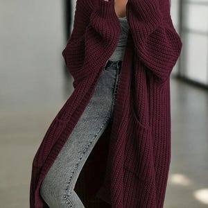 May include: A long, burgundy knit cardigan with two front pockets. The open-front sweater has a relaxed fit and long sleeves. The cardigan is made of a thick, textured knit material, perfect for layering. The garment is knee-length.