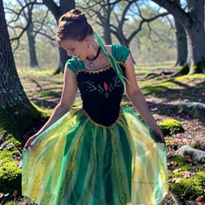 Puede incluir: Un vestido con un corpiño negro, escote con ribete dorado y un estampado floral colorido. La falda está hecha de capas de tela verde y amarilla semitransparente con un diseño estampado. Se lleva un collar tipo gargantilla negro.