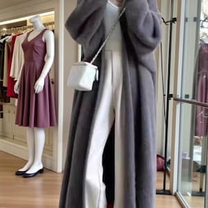 May include: A long, gray, faux fur coat worn over white pants and a white top. A small, white, square purse with a silver chain strap hangs from the shoulder. A mannequin in a burgundy dress is visible in the background.