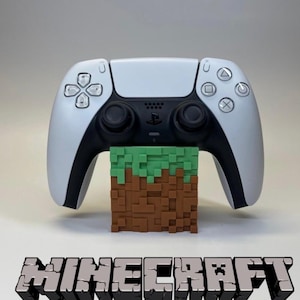 May include: A white and black PlayStation 5 controller supported by a Minecraft-themed block. The block is brown and green, with the word "MINECRAFT" in a 3D font below the controller.