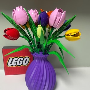 May include: A colourful bouquet of LEGO tulips in a purple vase. The flowers are various colours, including pink, red, yellow, and purple, with green stems and leaves. A red LEGO box is in the background.