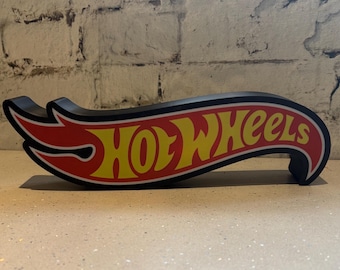 Może przedstawiać: Czarno-czerwony znak z logo Hot Wheels z żółtym wzorem płomienia. Znak ma kształt zakrzywionego skrzydła.