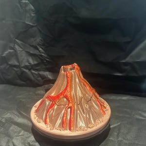 May include: A small, brown and red volcano model with a textured surface. The volcano has a crater at the top and red lava flowing down the sides. The volcano is sitting on a brown base.