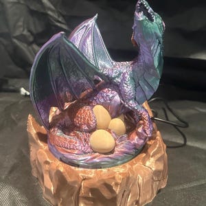 May include: A purple and green iridescent dragon figurine with wings spread, sitting on a brown rock base with eggs. The dragon is looking up with its mouth open.