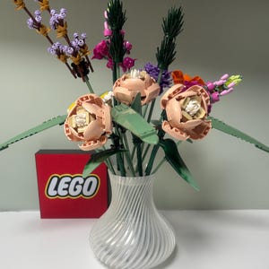 May include: A display of artificial flowers in a clear, ribbed glass vase. The arrangement features peach-toned flowers, purple lavender, and green foliage. A red box with the LEGO logo is visible in the background.