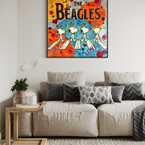 May include: A framed pop art print featuring Snoopy and friends crossing a street, with the words "THE BEAGLES" in bold letters. The artwork has a colorful background with orange, blue, and red accents. The print is displayed above a beige sofa with decorative pillows.
