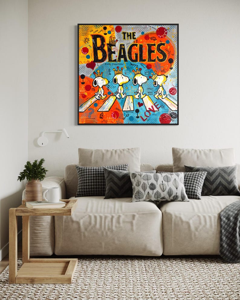 May include: A framed pop art print featuring Snoopy and friends crossing a street, with the words "THE BEAGLES" in bold letters. The artwork has a colorful background with orange, blue, and red accents. The print is displayed above a beige sofa with decorative pillows.