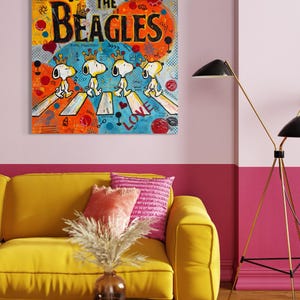 May include: A colorful pop art painting titled "The Beagles" features Snoopy characters crossing a street. The artwork has a vibrant palette of orange, yellow, blue, and red. The painting is displayed above a yellow sofa with pink pillows.