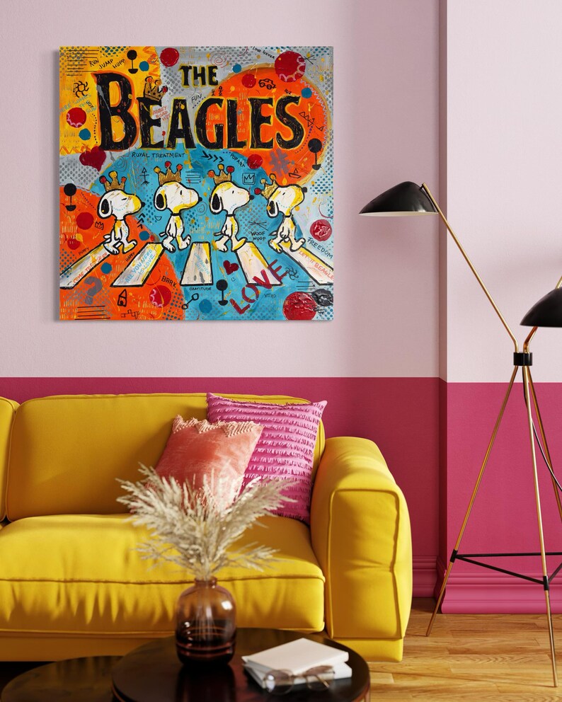 May include: A colorful pop art painting titled "The Beagles" features Snoopy characters crossing a street. The artwork has a vibrant palette of orange, yellow, blue, and red. The painting is displayed above a yellow sofa with pink pillows.