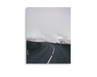May include: A photograph of a winding asphalt road with a white centre line, leading into a misty, overcast landscape. Yellow posts line the road's edge. The sky is filled with a thick, grey fog.