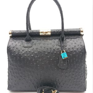 May include: A black handbag with a textured ostrich-style finish, featuring two top handles, gold-tone hardware, a central turn-lock closure, and a small bright blue padlock hanging from a strap. A detachable black shoulder strap is coiled at the base.