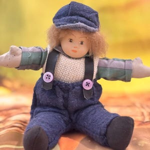 May include: A small doll with light brown hair wearing a blue cap, a beige knit sweater, and dark blue denim overalls with black straps and two light purple buttons. The doll has a pale face, painted features, and grey striped sleeves.