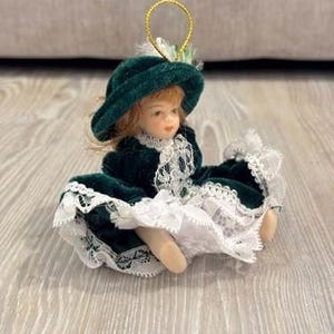 May include: A small porcelain doll with reddish-brown hair wearing a dark green velvet dress and a matching wide-brimmed hat. The dress features white lace trim along the collar and hem. A gold hanging loop is attached to the top of the hat.