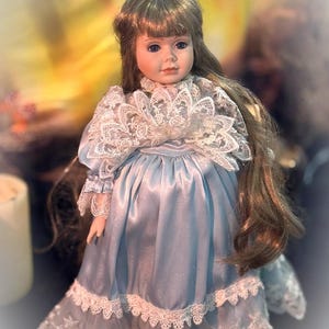 Peut inclure: Une poupée en porcelaine aux longs cheveux bruns et aux yeux bleus, vêtue d'une robe en satin bleu clair. La robe présente un col en dentelle blanche, des manches bordées de dentelle et un large volant en dentelle blanche avec broderie.