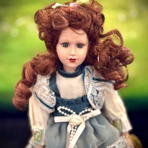 May include: A porcelain doll with curly auburn hair, blue eyes, and red lips. The doll wears a teal and white dress featuring a high collar, lace trim on the sleeves and hem, and a white ribbon sash with a central flower and pearl-like beads.
