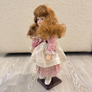 May include: A porcelain doll with auburn hair and blue eyes, dressed in a pink floral dress with a white apron and lace trim. The doll is mounted on a dark brown square base, exuding a vintage charm.