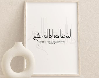 May include: A framed print with Arabic calligraphy in black, featuring the phrase "Guide us to the Straight Path" in English. A white, circular vase sits to the left. The print and vase are on a light-colored surface.