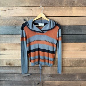 May include: A long-sleeved sweater with a cowl neck and drawstring hem. The sweater features horizontal stripes in orange, gray, and black. The sleeves and cowl neck are gray with black trim. The sweater is hanging on a wooden hanger.