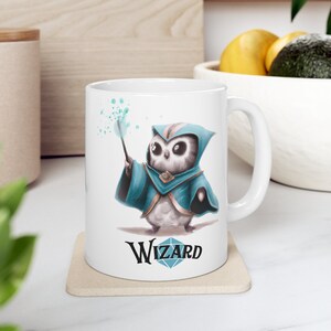 Könnte beinhalten: Eine weiße Keramiktasse mit einer Cartoon-Illustration einer Eule, die eine blaue Zaubererrobe trägt und einen Zauberstab hält. Die Eule zaubert mit grünen Funken. Der Text "WIZARD" ist in Schwarz unter der Eule mit einer Diamantenform gedruckt.