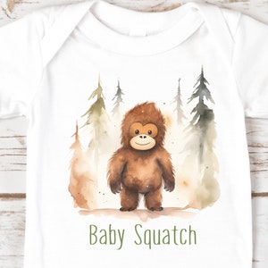 Może przedstawiać: Biały śpioszek dla niemowlęcia z akwarelową ilustracją brązowego Bigfoota stojącego w lesie z zielonymi drzewami. Tekst "Baby Squatch" jest nadrukowany na zielono pod ilustracją.
