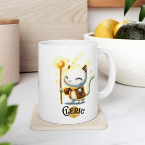 May include: White ceramic mug featuring a cartoon illustration of a white cat wearing a brown robe and holding a golden staff. The cat has a happy expression and is surrounded by yellow sparkles. The text "CLERIC" is printed below the cat in a stylised font.
