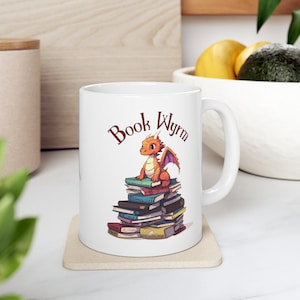 May include: White ceramic mug with a red and orange dragon sitting on a stack of books. The text "Book Wyrm" is printed on the mug.
