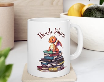 Op de afbeelding: Een witte keramische mok met een rode en oranje draak die op een stapel boeken zit. De tekst "Book Wyrm" staat op de mok gedrukt.