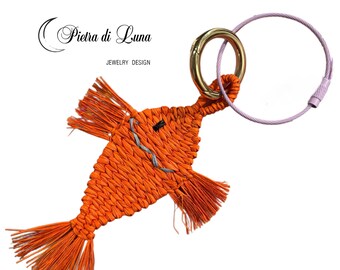 May include: An orange woven fish-shaped keychain with a gold-colored ring and a purple wire loop. The fish has fringed fins and tail, and a small gray stitched mouth. The text "Pietra di Luna" and "JEWELRY DESIGN" are in the upper left corner.