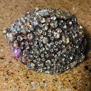 May include: A close-up of a large, round, jeweled brooch. The brooch is made of a dark red fabric base covered in hundreds of small, clear rhinestones. The rhinestones are arranged in a swirling pattern, creating a dazzling effect.