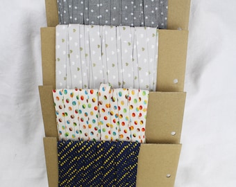 May include: Four rolls of fabric ribbon with different patterns. The ribbons are displayed on brown cardboard with holes punched in the side. The patterns include white polka dots on a gray background, white hearts on a beige background, colorful polka dots on a white background, and yellow circles on a navy blue background.