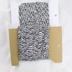 May include: Two rolls of black and white patterned fabric. The top roll has a polka dot pattern and the bottom roll has a zebra stripe pattern. The fabric is wrapped around a cardboard core and is displayed on a white background.