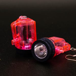 May include: Two pink plastic light-up earrings with a silver hook. The earrings are shaped like small lanterns with a clear plastic window that shows the light inside.