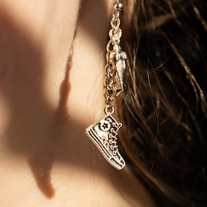 May include: Silver dangle earrings featuring a charm of a sneaker with a star on the side, a small charm of a bird, and a chain.