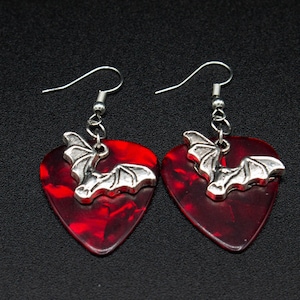 May include: A pair of red guitar pick earrings with silver bat charms. The earrings are on a black background.