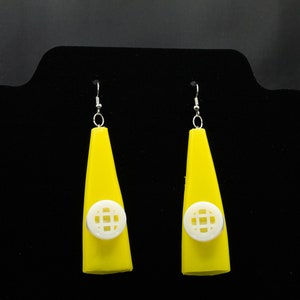 May include: A pair of yellow dangle earrings with white geometric designs. The earrings are on a black display stand.