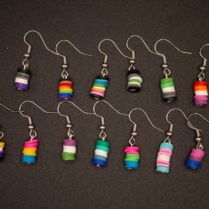 May include: Thirteen individual dangle earrings featuring small, cylindrical clay beads arranged in various multi-colored striped patterns. Each earring is attached to a silver-toned metal hook, showcasing a variety of distinct color combinations.
