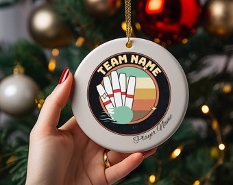 May include: A round, white ceramic ornament with a bowling theme. The design features bowling pins, a bowling ball, and text that reads "Team Name" and "Player Name" in a retro style. The ornament hangs from a gold-colored string.