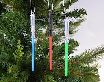 May include: Three lightsaber Christmas tree ornaments with silver handles and colored blades: blue, red, and green. The ornaments hang from white strings against a backdrop of a green Christmas tree.