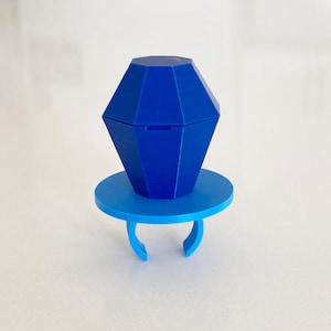 May include: A blue plastic ring with a blue diamond-shaped box on top. The ring is designed to hold a small item, such as a ring or a piece of jewelry.