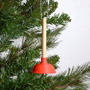 Op de afbeelding: Een nieuwigheid kerstboomversiering in de vorm van een miniatuur ontstopper. De ontstopper heeft een felrode zuignap en een lichtbruine handgreep. Hij hangt aan een zilveren ketting, tegen een achtergrond van groene kerstboomtakken.