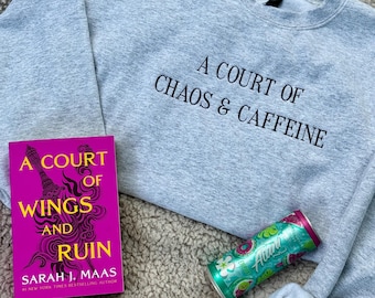 May include: A light gray sweatshirt with the text "A Court of Chaos & Caffeine" in black. A book titled "A Court of Wings and Ruin" and a can of Alani Nu energy drink are also present, along with a pair of black glasses.
